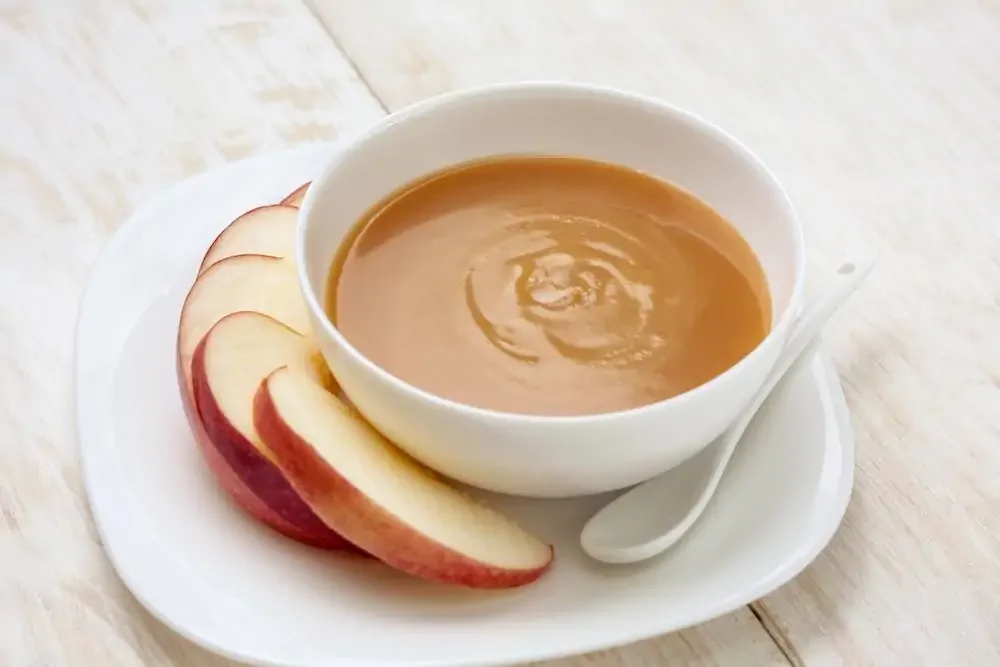 white bowl of caramel sauce with sliced apple and a spoon on the side