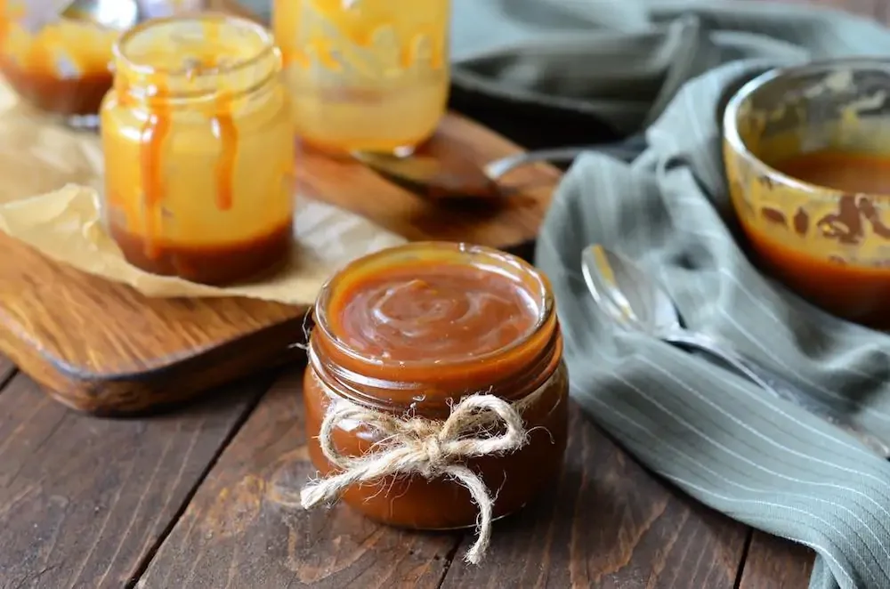 jar of homemade vegan caramel sauce on a wooden table with blue napkins, spoons, and other jars