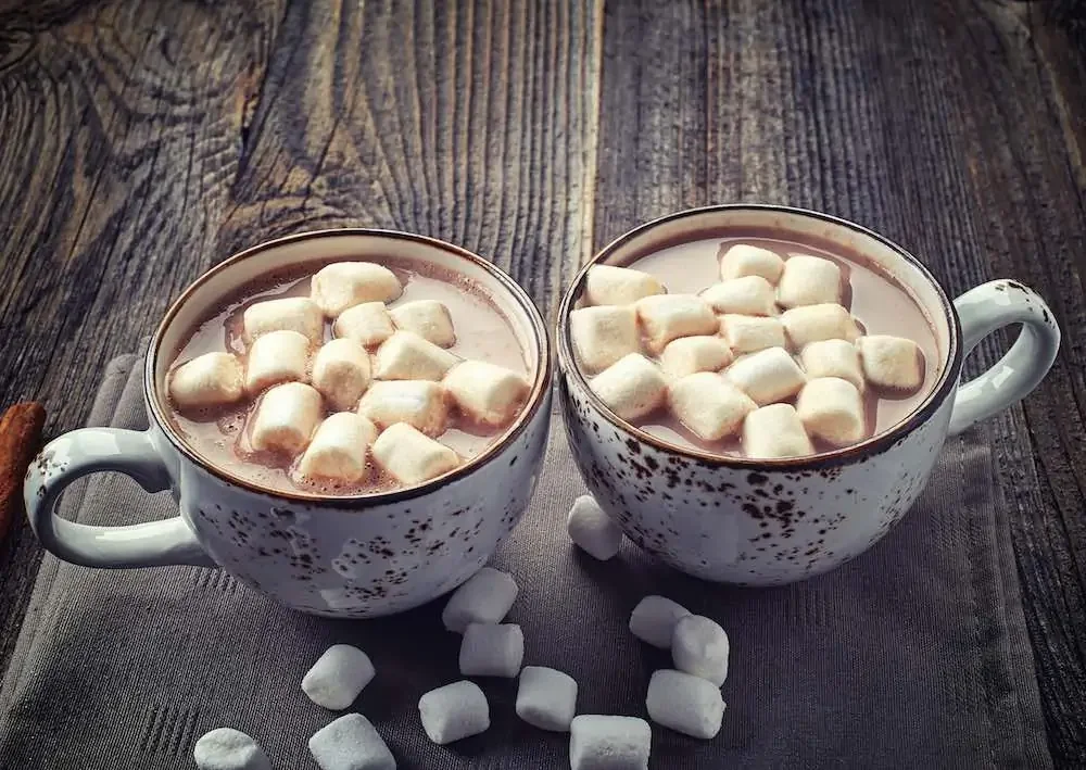 two cups of hot chocolate with vegan marshmallows on top and scattered around on a wooden table